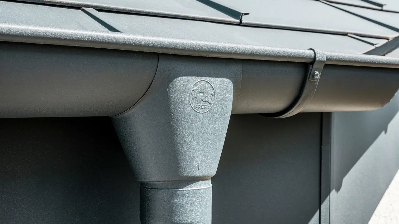 Nahaufnahme eines Hausdaches mit PREFA Dachplatte R.16 in Anthrazit sowie detailreicher Ansicht der farblich abgestimmten PREFA Dachrinne und Fallrohr aus Aluminium bei sonnigem Wetter.