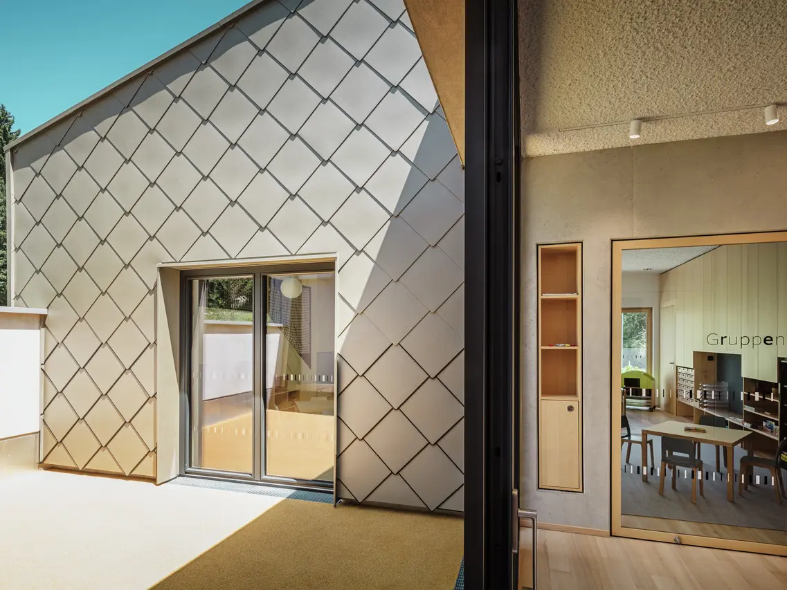 Edificio moderno con tetto e rivestimento di facciata PREFA composto da elementi romboidali 44 x 44, fotografato alla luce del giorno. L'edificio mostra un'armoniosa integrazione di elementi architettonici tradizionali e moderni, con i rombi metallici come elementi di spicco. Le ampie vetrate offrono una vista su un interno luminoso, la cosiddetta sala gruppi, arredata con tavoli e sedie per riunioni o lezioni.  