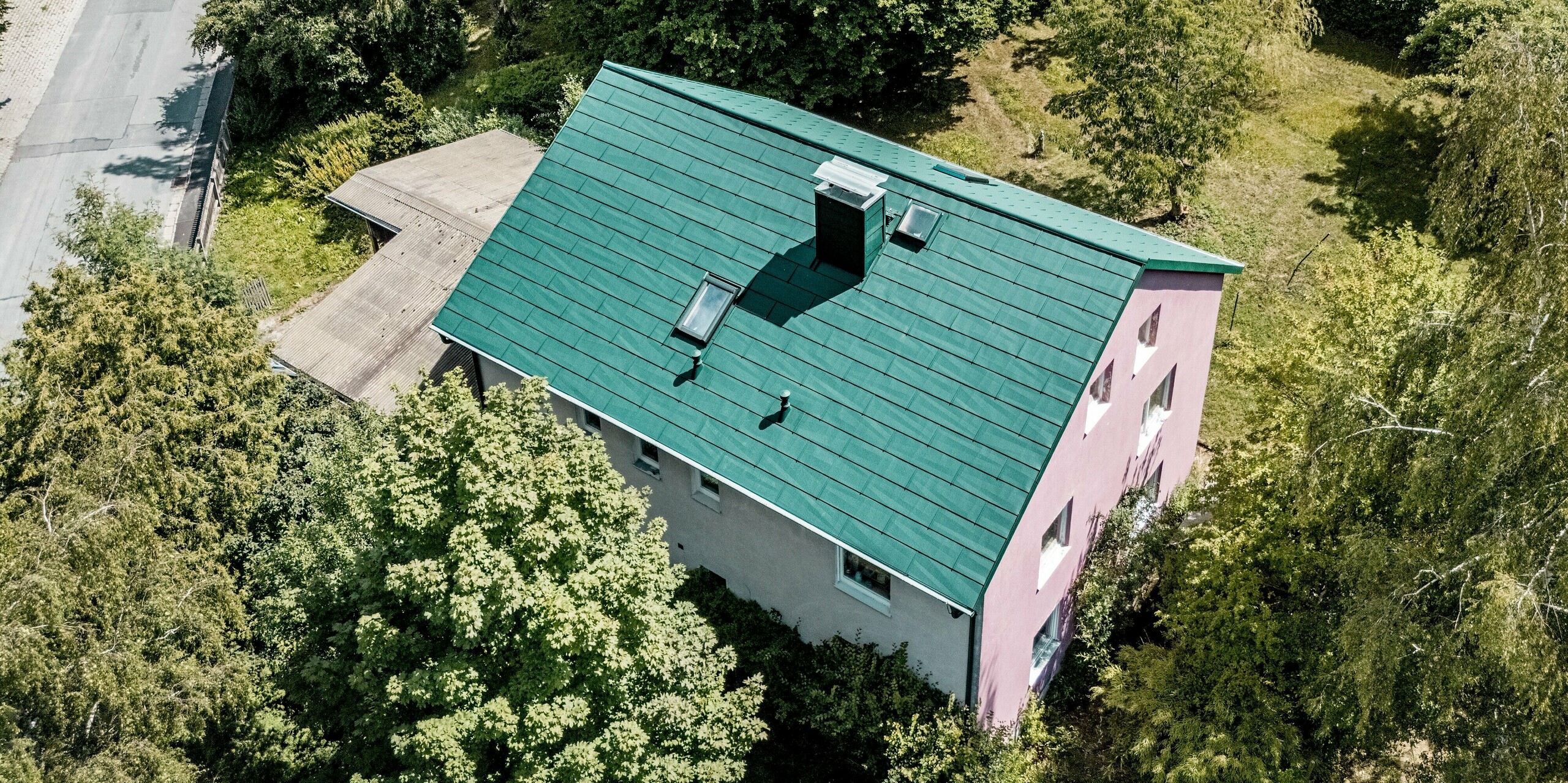 Vue aérienne d'une maison individuelle rénovée à Schwarzenbach-Martinlamitz, dotée de panneaux de toiture PREFA FX.12 couleur Vert Mousse P.10. La toiture moderne en aluminium séduit par ses lignes épurées, son puits de lumière intégré et sa cheminée élégamment habillée. Le contraste entre le vert mousse de la toiture et le violet de la façade à pignon est particulièrement saisissant. Entourée d'une végétation luxuriante, la maison s'intègre harmonieusement au paysage. La toiture PREFA offre une durabilité exceptionnelle, une résistance aux intempéries et une surface sans entretien – idéale pour les rénovations durables.