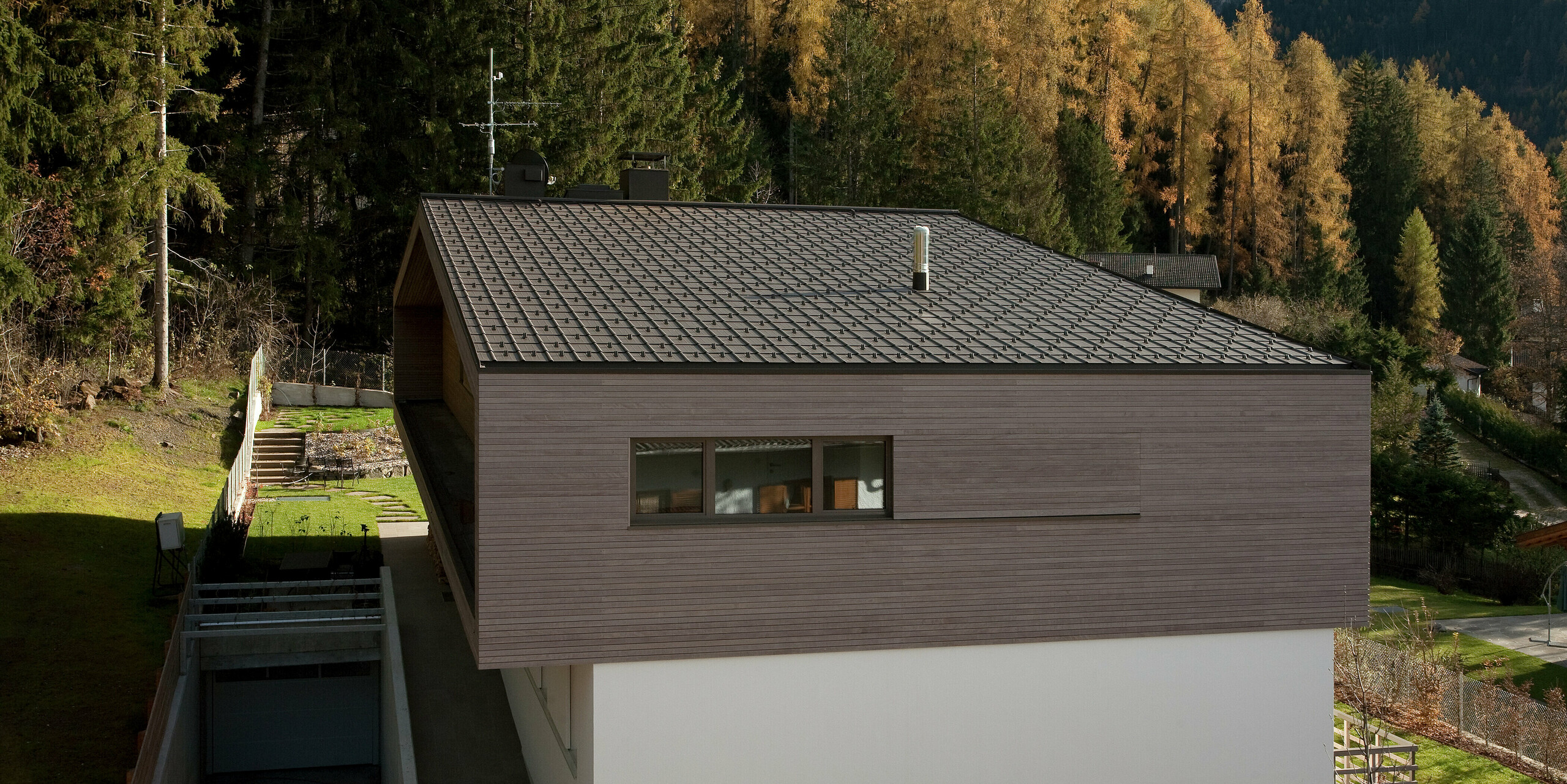 Moderne Architektur mit PREFA Dachplatten in P.10 Braun, umgeben von einem naturnahen Garten und Waldlandschaft. Der minimalistische Baukörper mit Holzfassade fügt sich harmonisch in die Umgebung ein.
