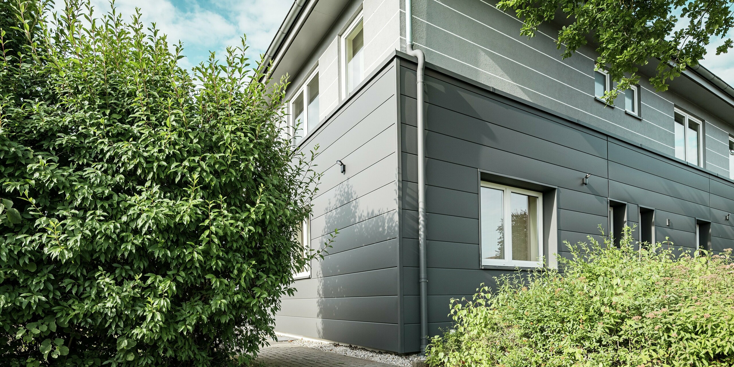 Les Sidings PREFA P.10 Gris sombre habillent la façade de la Guilde des ramoneurs de Bindlach, lui conférant une allure moderne et épurée. Ces panneaux en aluminium, outre leur esthétique, offrent une protection durable contre les intempéries. L'image illustre l'harmonie entre la simplicité de la façade et la végétation au premier plan. Durables et nécessitant peu d'entretien, ces panneaux constituent une solution écologique pour la conception de façades modernes.