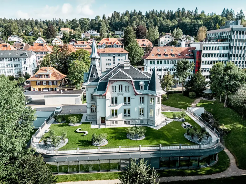 Weitläufige Gesamtansicht der Manufacture Horlogère du Locle in der Schweiz, eingebettet in eine grüne Parklandschaft und umgeben von historischer Bebauung. Im Fokus steht die ikonische Dacharchitektur mit eleganten Türmen, steilen Flächen und geschwungenen Formen. PREFA Dachschindeln in P.10 Schwarz betonen präzise die Geometrie, während PREFALZ in Patinagrau fließende Übergänge ermöglicht. Das leichte Aluminiumdach überzeugt durch Langlebigkeit, Witterungsbeständigkeit und maximale Gestaltungsfreiheit bei komplexen Dachformen.