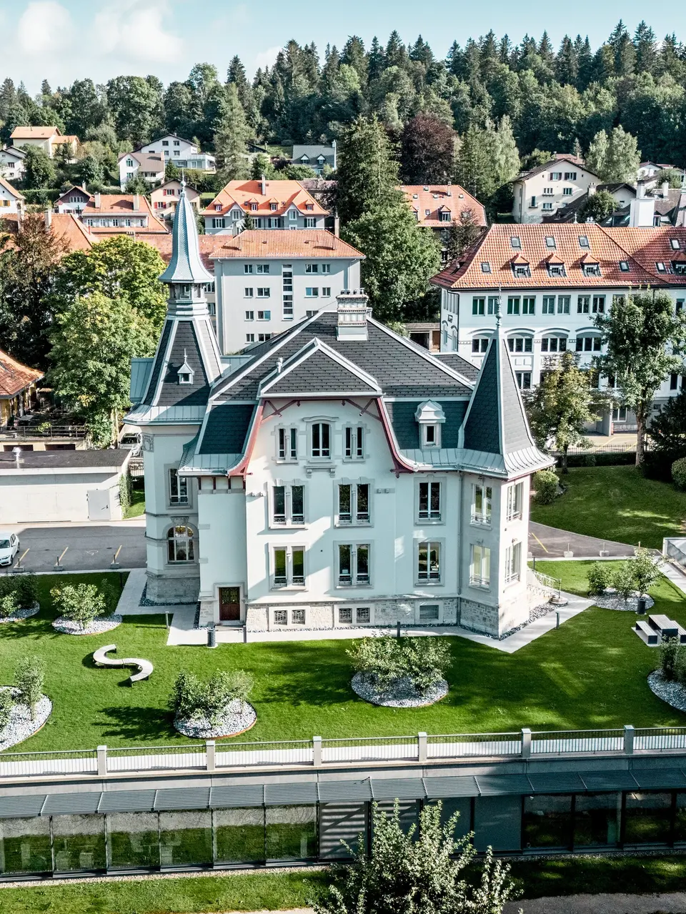 Weitläufige Gesamtansicht der Manufacture Horlogère du Locle in der Schweiz, eingebettet in eine grüne Parklandschaft und umgeben von historischer Bebauung. Im Fokus steht die ikonische Dacharchitektur mit eleganten Türmen, steilen Flächen und geschwungenen Formen. PREFA Dachschindeln in P.10 Schwarz betonen präzise die Geometrie, während PREFALZ in Patinagrau fließende Übergänge ermöglicht. Das leichte Aluminiumdach überzeugt durch Langlebigkeit, Witterungsbeständigkeit und maximale Gestaltungsfreiheit bei komplexen Dachformen.