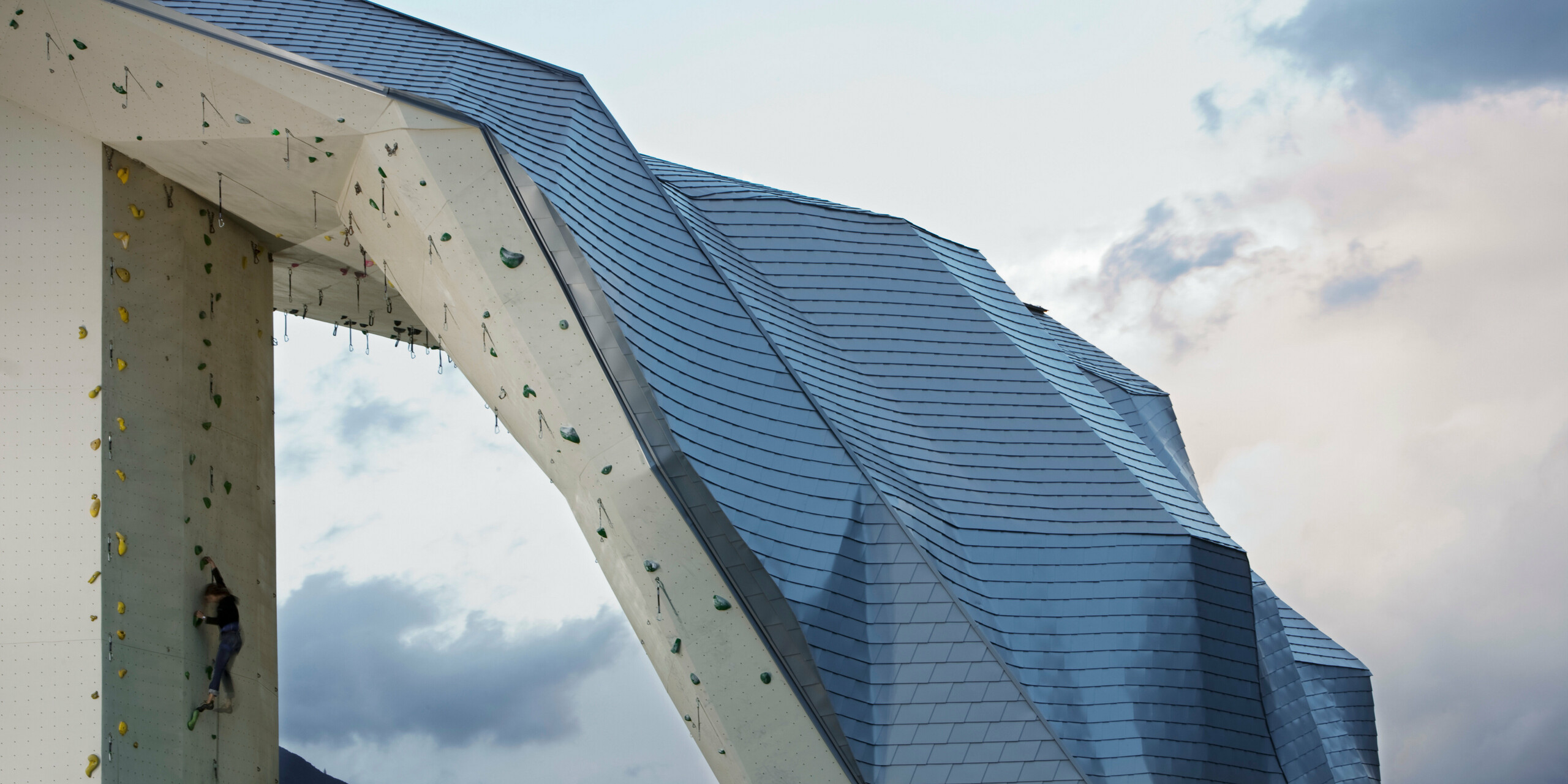 Im Vordergrund die Spitze des Kletterturms Imst mit Fassade bzw. Dach aus PREFA Dachschindeln in Silbermetallic. Am linken Bildrand eine Kletterin, am rechten Bildrand ein schöner Himmel in abendlicher Stimmung.