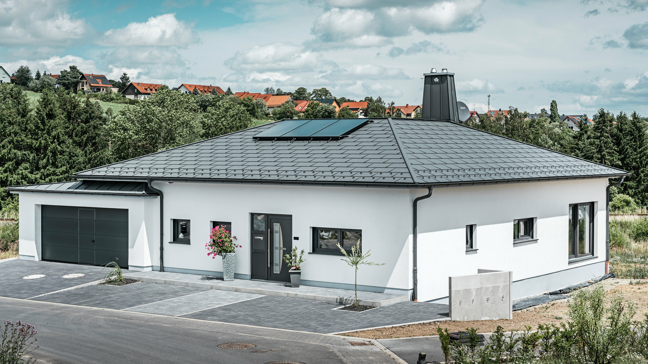 Elegantes Einfamilienhaus in Ilmenau, Thüringen, mit einem hochwertigen PREFA Dach in der Farbe P.10 Dunkelgrau, ausgestattet mit der robusten Dachplatte R.16. Die Garage harmoniert in Farbe und Stil durch die Verwendung von PREFALZ. Der Blickfang des Anwesens ist das funktional gestaltete Atrium und der mit dem PREFA Stier verzierte Schornstein, welcher das moderne Design und die innovative Dachtechnologie von PREFA unterstreichen. Die Solarpaneele fügen sich nahtlos in das nachhaltige Konzept des Hauses ein. Abgerundet wird das durchdachte Gesamtkonzept des Bungalows durch das PREFA Dachentwässerungssytem, welches ebenfalls in Dunkelgrau gehalten ist.