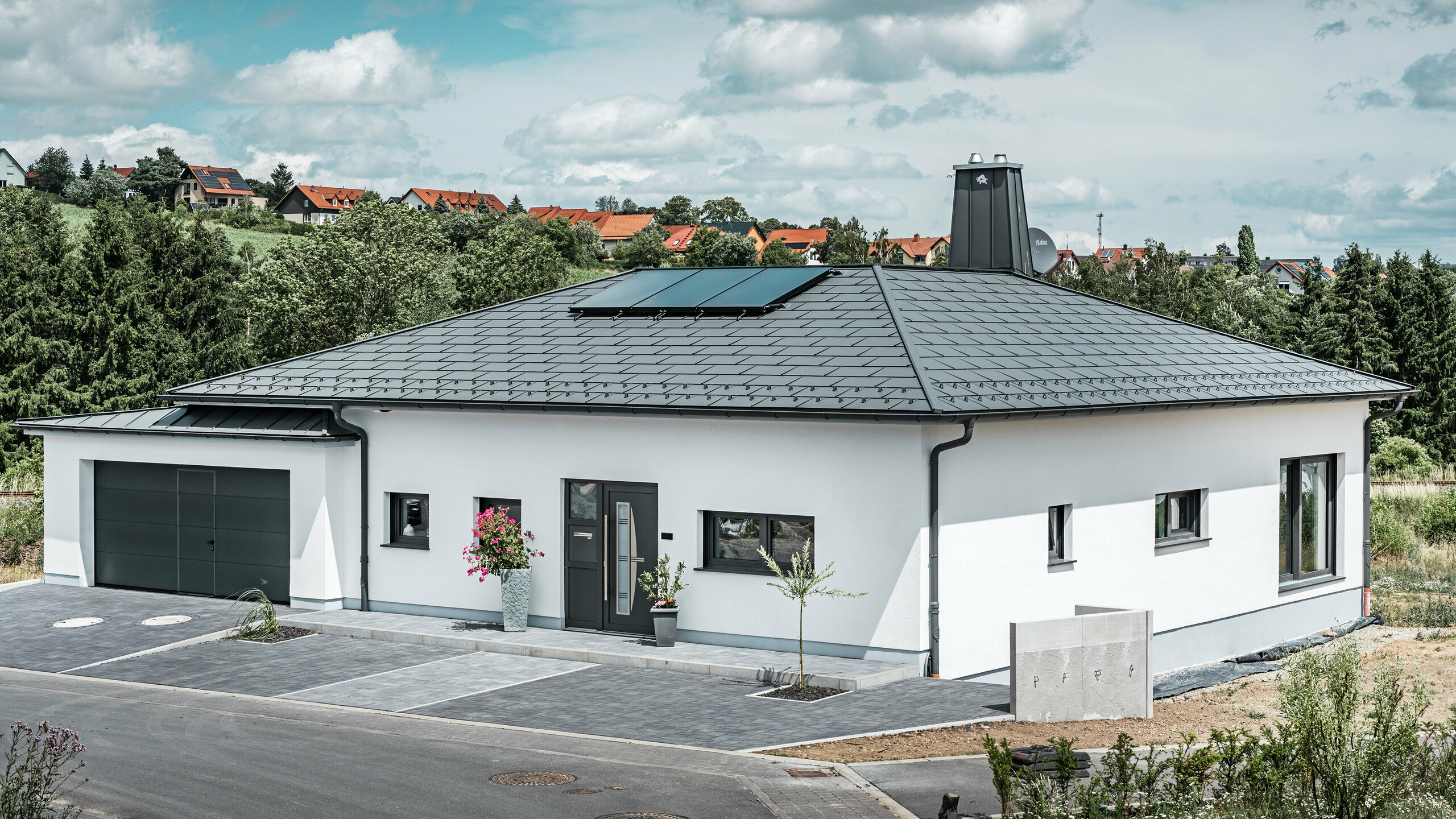 Elegantes Einfamilienhaus in Ilmenau, Thüringen, mit einem hochwertigen PREFA Dach in der Farbe P.10 Dunkelgrau, ausgestattet mit der robusten Dachplatte R.16. Die Garage harmoniert in Farbe und Stil durch die Verwendung von PREFALZ. Der Blickfang des Anwesens ist das funktional gestaltete Atrium und der mit dem PREFA Stier verzierte Schornstein, welcher das moderne Design und die innovative Dachtechnologie von PREFA unterstreichen. Die Solarpaneele fügen sich nahtlos in das nachhaltige Konzept des Hauses ein. Abgerundet wird das durchdachte Gesamtkonzept des Bungalows durch das PREFA Dachentwässerungssytem, welches ebenfalls in Dunkelgrau gehalten ist.