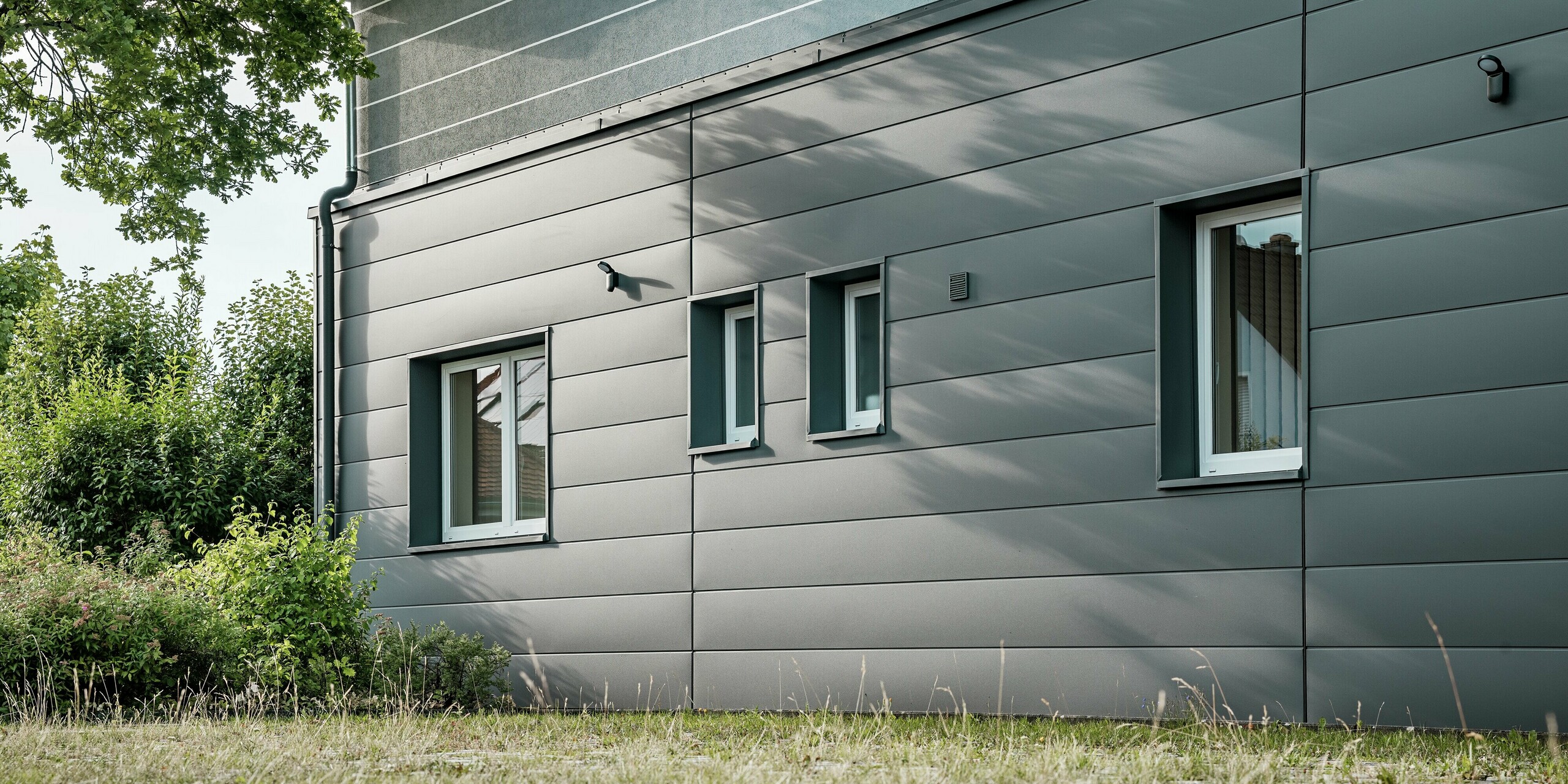 L'image présente un détail de la façade modernisée de la guilde des ramoneurs de Bindlach, revêtue de Siding PREFA couleur Gris sombre P.10. La structure apparente des panneaux d'aluminium confère au bâtiment une allure à la fois professionnelle et esthétique. Le jeu d'ombres harmonieux, qui souligne l'interaction entre la lumière et l'architecture, est particulièrement saisissant. Grâce à la durabilité et à la facilité d'entretien du bardage PREFA, la façade conserve son excellent état durablement.