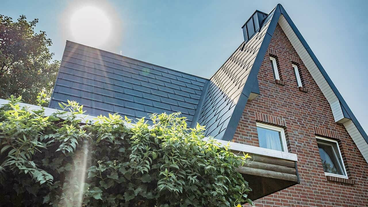 Toit à deux pans recouvert avec la sobre tuile en aluminium R.16 PREFA couleur anthracite. La façade est une façade en brique rustique, le soleil se lève juste derrière la maison.