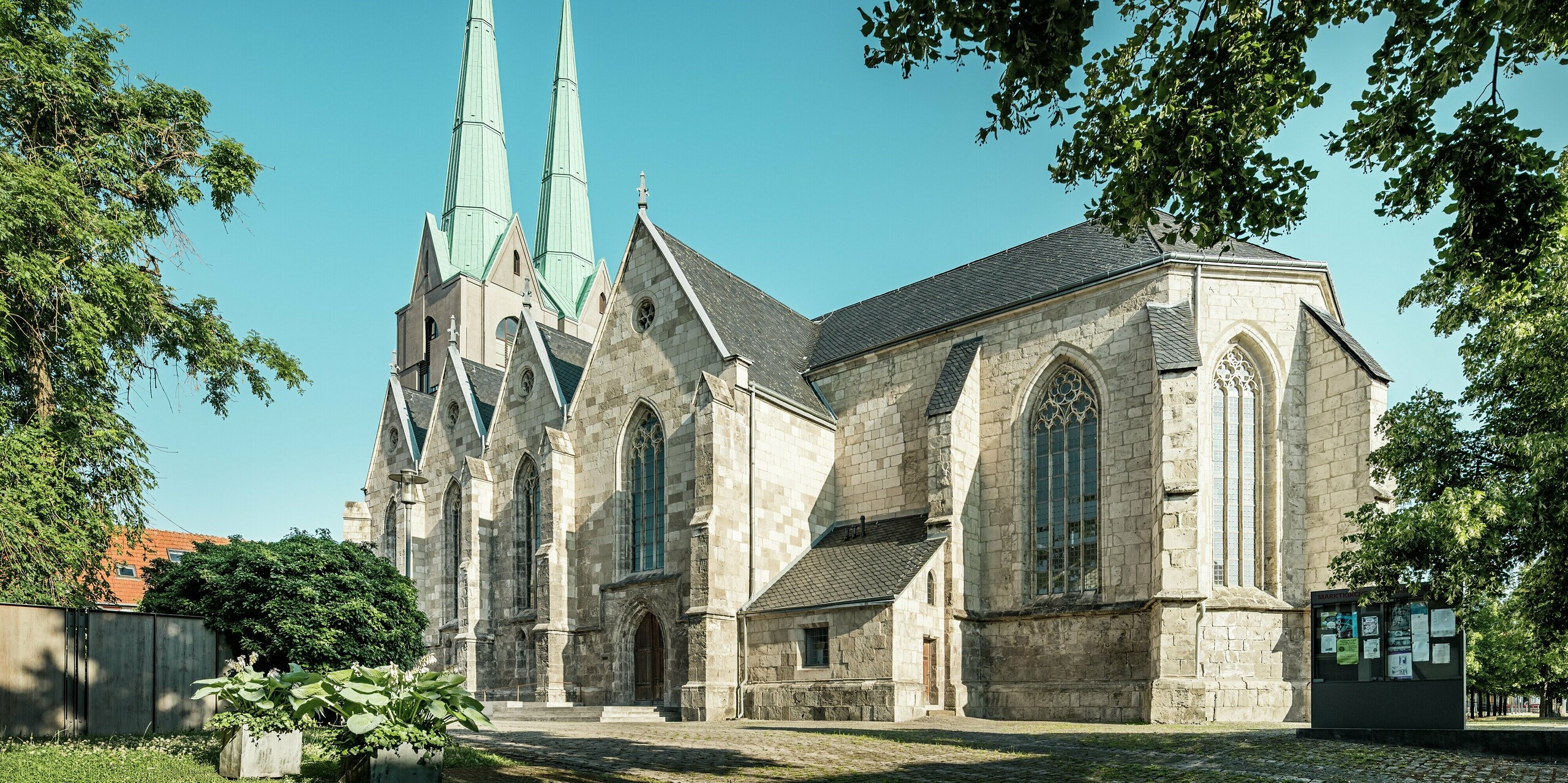 Vue latérale harmonieuse de l'église Saint-Jean rénovée d'Ellrich, encadrée par des arbres et un ciel d'un bleu éclatant. Les flèches remarquables, revêtues de PREFALZ en vert de gris P.10, contrastent fortement avec la maçonnerie historique en pierre naturelle. La toiture en aluminium de haute qualité souligne la modernité de l'édifice et offre une durabilité et une protection optimales.