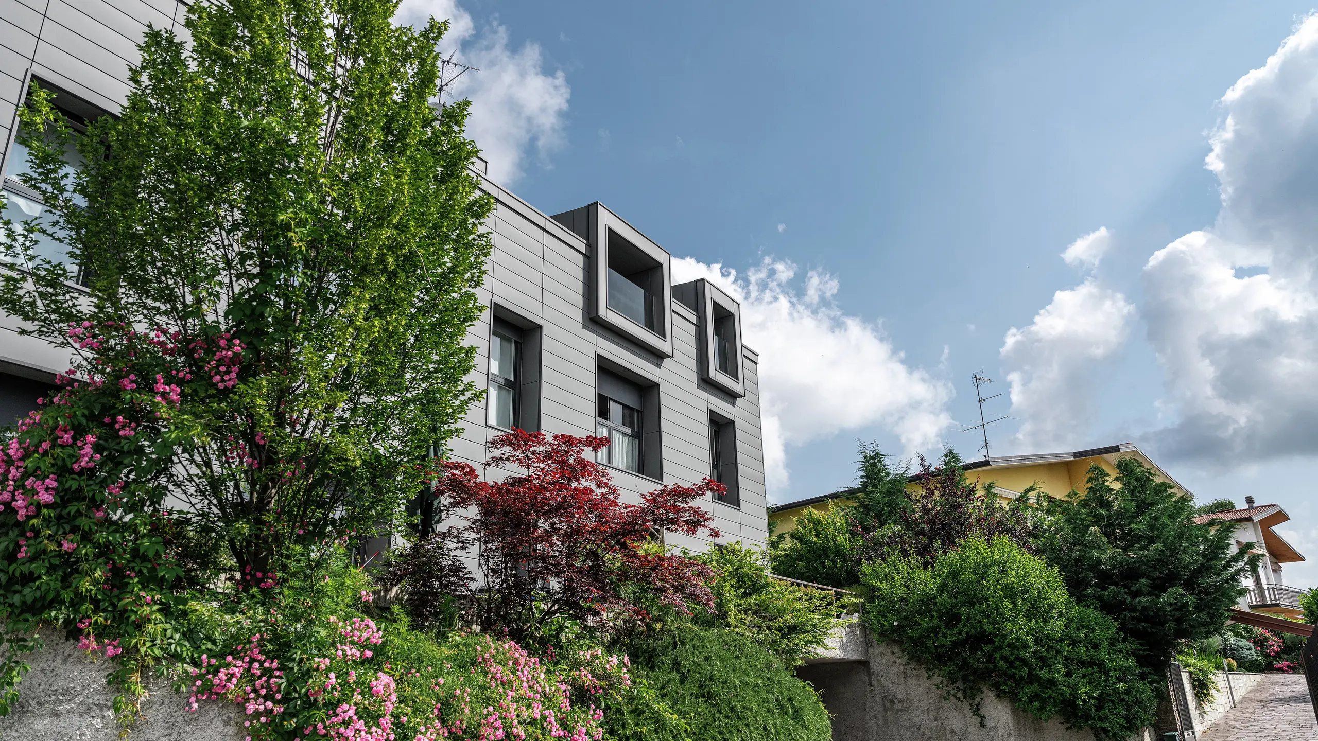 Complexe résidentiel moderne à Faloppio, en Italie, avec une façade élégante en Sidings PREFA. Les fenêtres en porte-à-faux offrent un attrait architectural unique, tandis que les plantes et les fleurs environnantes rehaussent la beauté naturelle de la propriété. Le revêtement en aluminium durable de PREFA offre une solution esthétique et fonctionnelle dans toutes les conditions météorologiques.