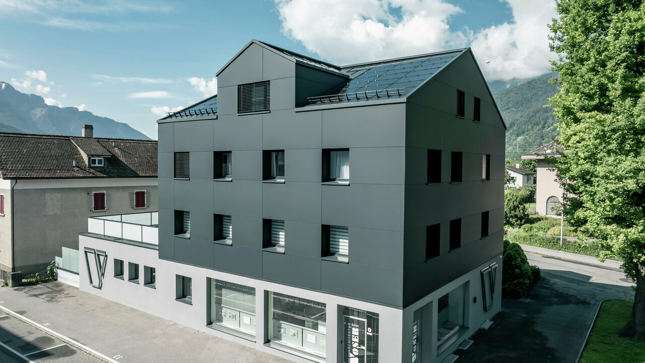 Modernisiertes Wohn- und Geschäftsgebäude nach der Fassadensanierung mit PREFABOND Aluminium Verbundplatten in Anthrazit. Die klare, elegante Linienführung der neuen Fassade verleiht dem Gebäude ein modernes Erscheinungsbild. Im Erdgeschoss befindet sich das Architekturbüro Voser. Die Umgebung mit Bäumen und Bergen rundet die harmonische Optik ab.