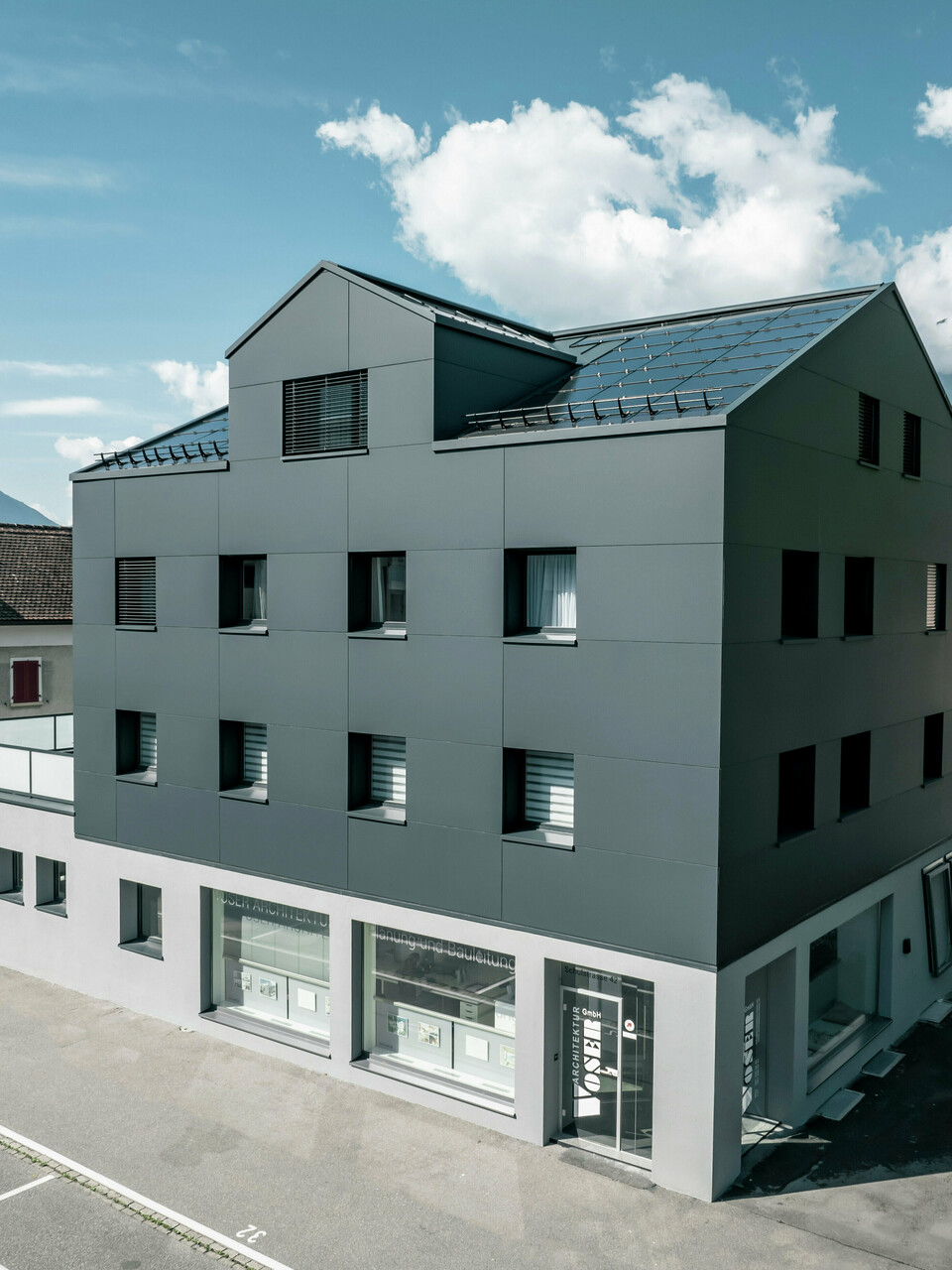 Modernisiertes Wohn- und Geschäftsgebäude nach der Fassadensanierung mit PREFABOND Aluminium Verbundplatten in Anthrazit. Die klare, elegante Linienführung der neuen Fassade verleiht dem Gebäude ein modernes Erscheinungsbild. Im Erdgeschoss befindet sich das Architekturbüro Voser. Die Umgebung mit Bäumen und Bergen rundet die harmonische Optik ab.