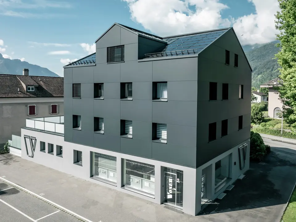 Modernisiertes Wohn- und Geschäftsgebäude nach der Fassadensanierung mit PREFABOND Aluminium Verbundplatten in Anthrazit. Die klare, elegante Linienführung der neuen Fassade verleiht dem Gebäude ein modernes Erscheinungsbild. Im Erdgeschoss befindet sich das Architekturbüro Voser. Die Umgebung mit Bäumen und Bergen rundet die harmonische Optik ab.