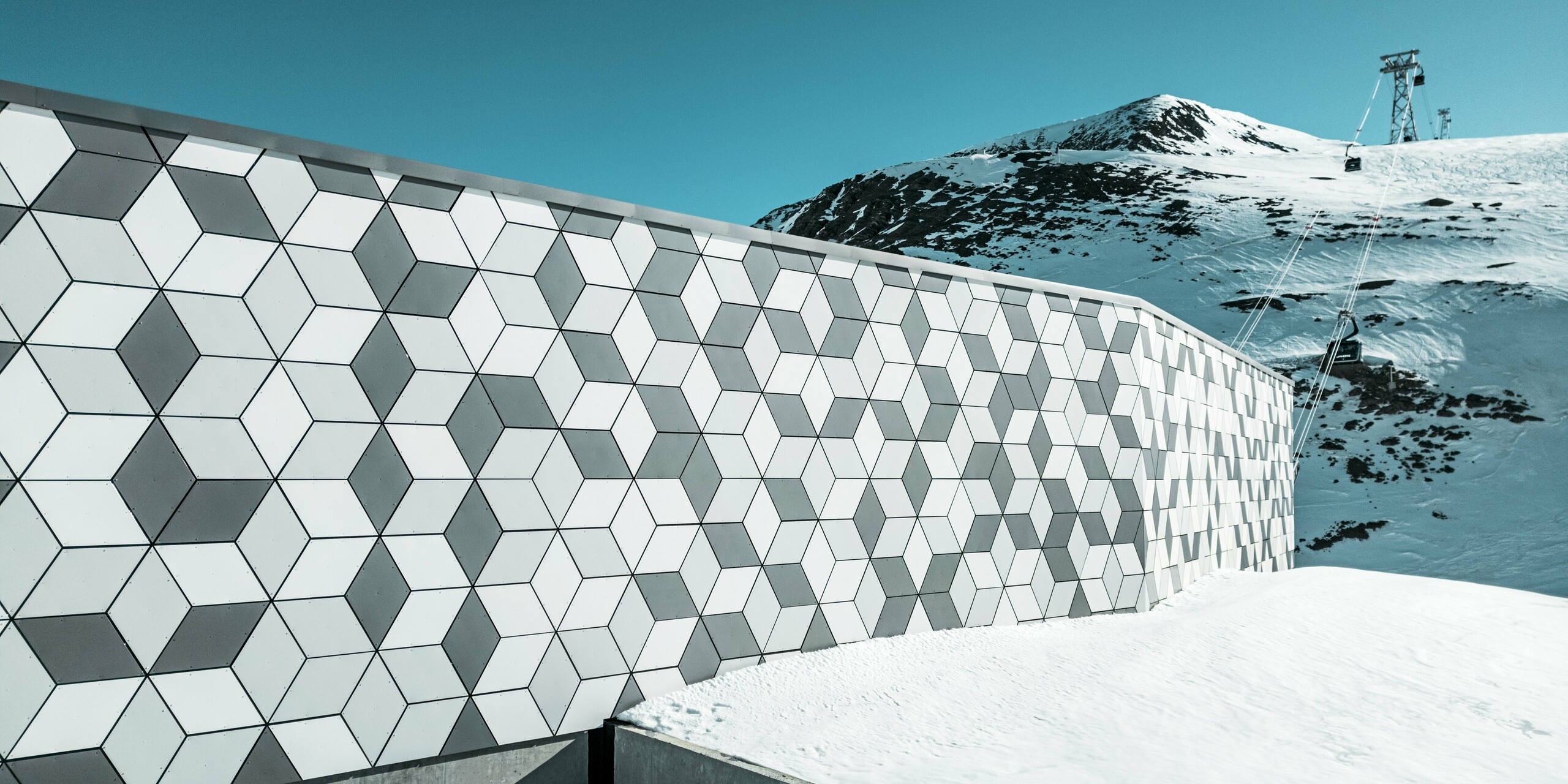Die Fassade der Seilbahnstation in Les Deux Alpes ist ein Meisterwerk moderner Architektur. Sie besteht aus PREFABOND Aluminium Verbundplatten, die in einem auffälligen Rautenmuster aus Weiß und Grau angeordnet sind. Die geometrische Gestaltung fügt sich harmonisch in die schneebedeckte Berglandschaft ein und bietet dank der robusten und wetterbeständigen Materialien lang anhaltenden Schutz vor extremen Witterungsbedingungen. Der Blick vorbei an der Fassade zeigt den Weg der Gondel hinauf zum Berg.