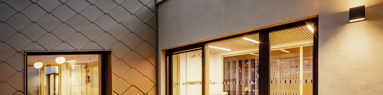 Vista serale di una parte del centro di formazione di Gerlos con una facciata artistica. Sul lato sinistro si trova la parete a rombi 44x44 di PREFA, mentre l'ingresso sul lato destro è un'ampia vetrata che conduce a un interno ben illuminato con sedie e tavoli. La calda illuminazione interna contrasta con la luce opaca del crepuscolo e sottolinea l'architettura moderna e il design della facciata.  