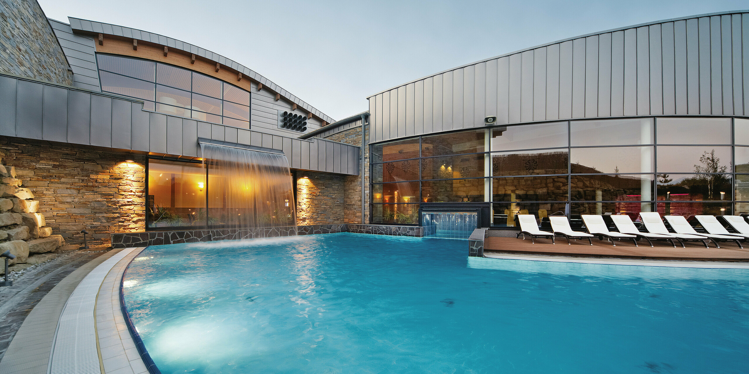 In der Dämmerung wird die Therme Białka durch die beleuchteten Fenster und die reflektierenden Wasserflächen besonders in Szene gesetzt. Die PREFALZ-Fassade in der Sonderfarbe Delphin unterstreicht die elegante Architektur, die durch die Kombination von Aluminium und Naturmaterialien eine harmonische und zeitlose Wirkung erzielt.