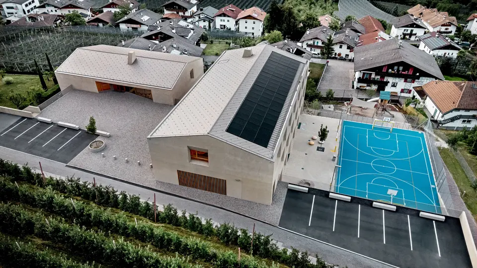 Zračna snimka obrazovnog centra Riffian u Južnom Tirolu s dječjim vrtićem, osnovnom školom i sportskim igralištem. PREFA krovni rombovi dimenzija 44 × 44 u boji P.10 bronca tvore preciznu i dugotrajnu aluminijsku krovnu oblogu koja se skladno uklapa u alpski krajolik. Moderna arhitektura ističe se otpornošću na vremenske uvjete i čistim dizajnom do najsitnijih detalja.