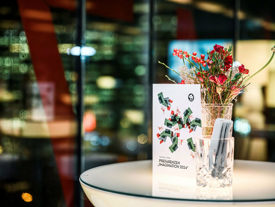Vue d'une invitation à l'événement PREFARENZEN 2024 posée sur une table devant un arrangement floral dans un pichet en verre, derrière lequel s'étend un arrière-plan flou.