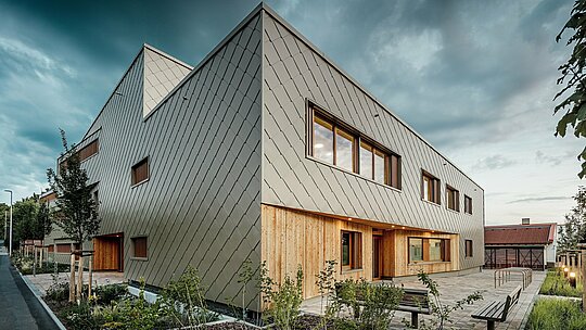In der sanften Abendstimmung entfaltet das Hybridgebäude der Kindertagesstätte in Alzenau seine volle architektonische Wirkung. Die markante Fassade aus bronzefarbenen PREFA Wandrauten 44 × 44 verleiht dem Gebäude eine elegante, wetterbeständige Hülle mit klarer Struktur. Im Erdgeschoss sorgen vertikale Holzverkleidungen für einen natürlichen, warmen Kontrast zum Aluminium und betonen den Eingangsbereich sowie die geschützten Aufenthaltszonen. Die reduzierte, moderne Formsprache harmoniert mit der gepflegten Außenanlage, in der Bänke, Fahrradständer und junge Bäume zum Verweilen einladen. Das Bild zeigt eindrucksvoll, wie langlebige PREFA Produkte zu einem nachhaltigen, funktionalen und zugleich einladenden Bildungsbau beitragen.