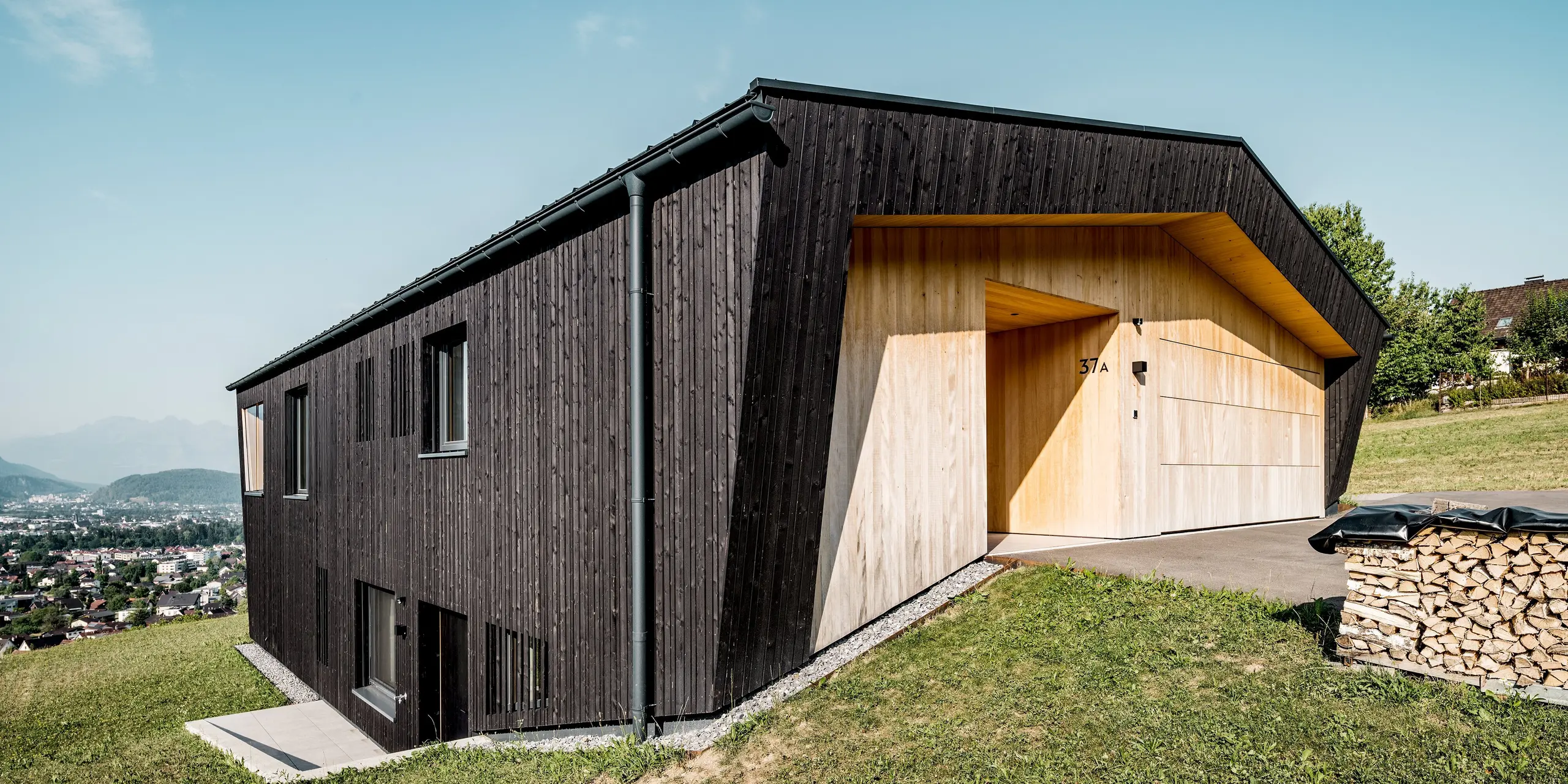 Das moderne Einfamilienhaus in Röthis begeistert mit seiner außergewöhnlichen Architektur: Die dunkle Holzfassade bildet einen eindrucksvollen Kontrast zum hellen Eingangs- und Garagenbereich. Das PREFALZ Dach in P.10 Anthrazit wird durch die farblich abgestimmte PREFA Dachentwässerung mit Hängerinne und Ablaufrohr ergänzt. Die langlebigen Aluminiumprodukte vereinen zuverlässigen Witterungsschutz mit zeitlosem Design.