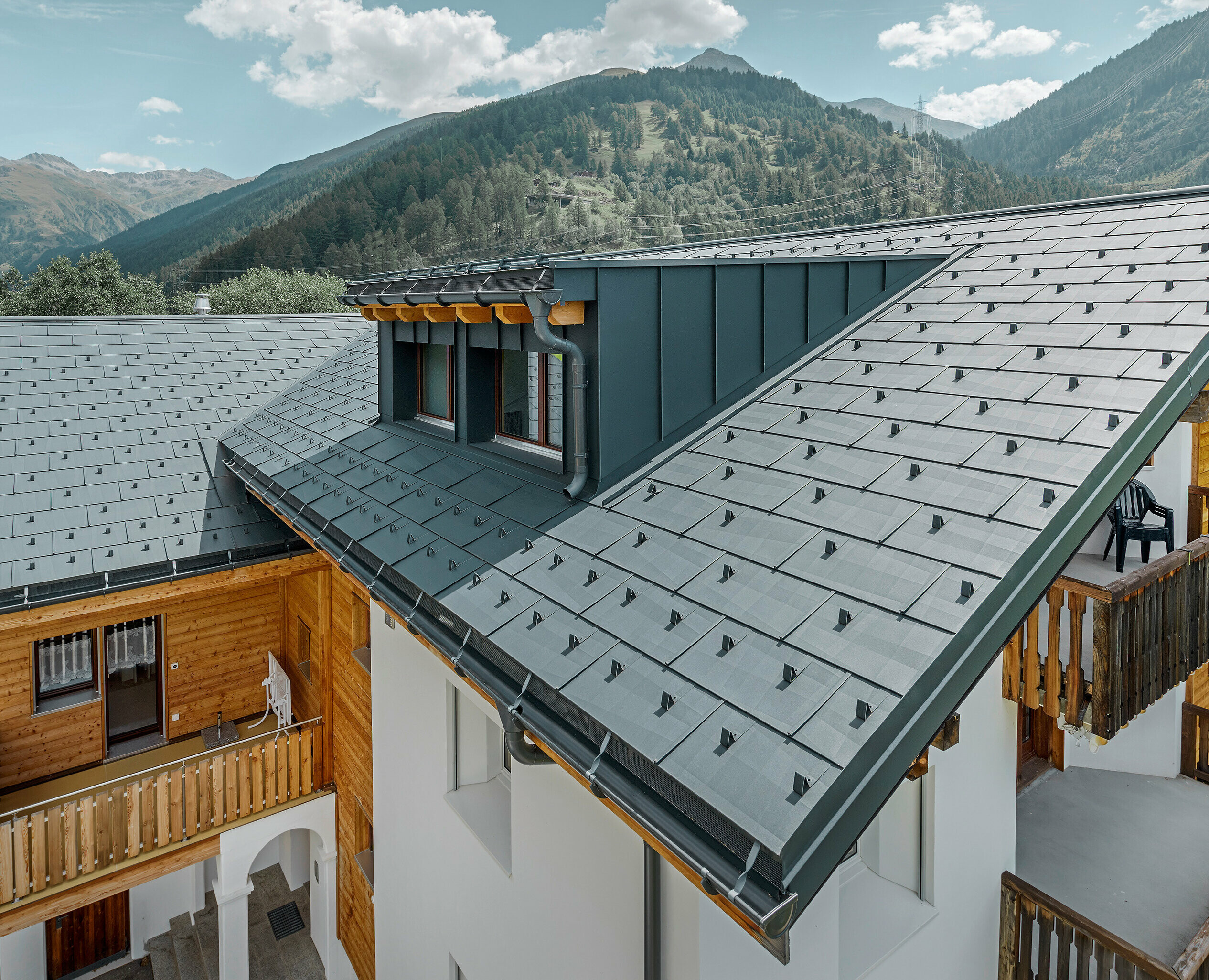 Immeuble d’habitation comportant une lucarne et un toit à deux pans recouvert de panneaux de toit FX.12 PREFA couleur anthracite. On aperçoit des montagnes en arrière-plan.