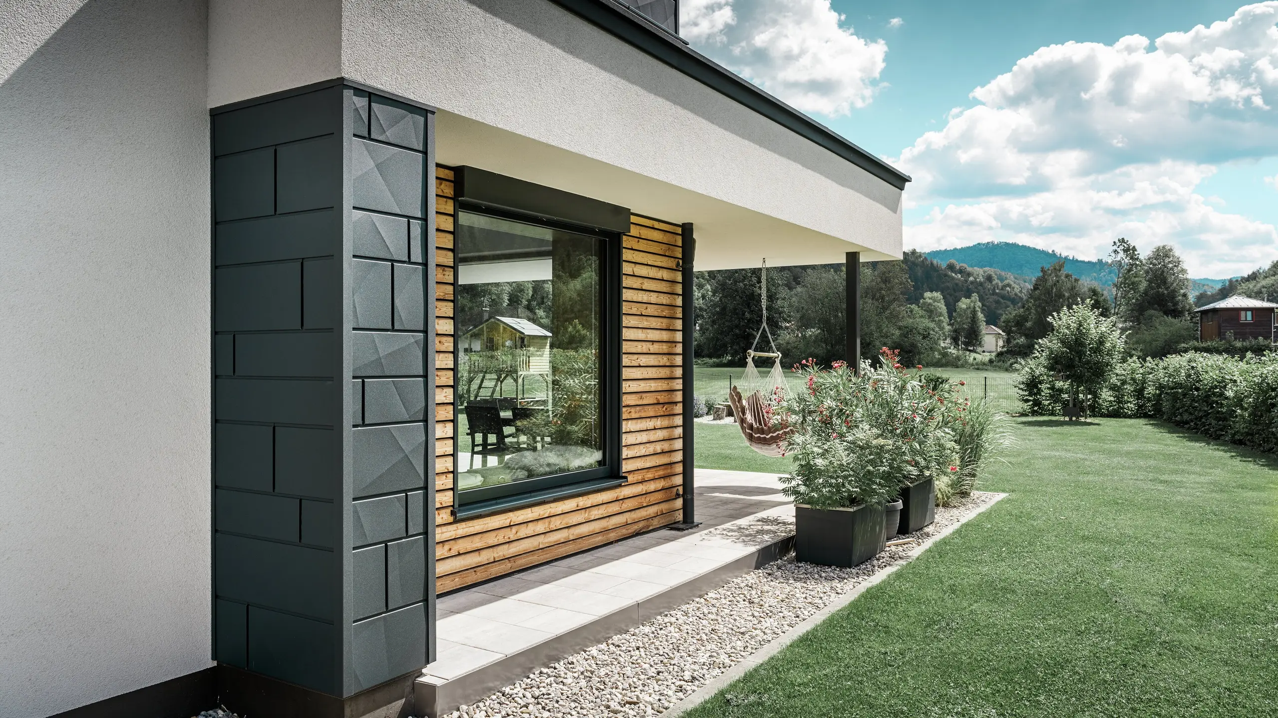 Particolare di una casa unifamiliare di nuova costruzione. Qui sono stati utilizzati diversi materiali per la facciata. La facciata intonacata di bianco è messa in risalto dalla Doga.X in alluminio  di colore antracite e dai pannelli orizzontali in legno che racchiudono una grande finestra. Sulla terrazza è appesa una poltrona pensile.