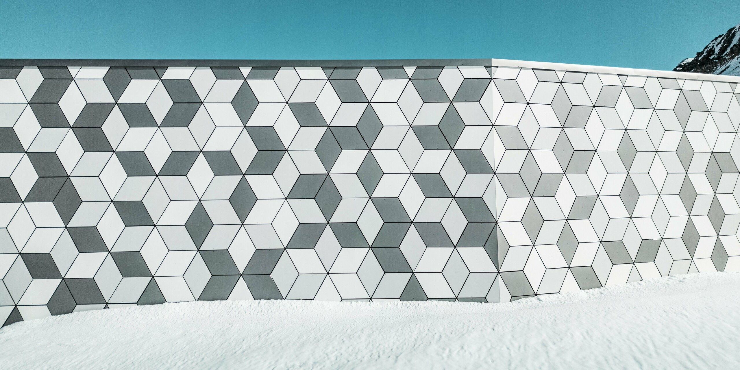 Die Nahaufnahme der Fassade der Seilbahnstation Jandri in Les Deux Alpes zeigt die präzise Anordnung der Rauten aus PREFABOND Aluminium Verbundplatten. Die angefertigten Rauten sind in einem auffälligen Muster angeordnet. Durch die harmonische Farbgestaltung in unterschiedlichen Weiß- und Grautönen fügt sich die Fassade perfekt in die schneebedeckte Alpenlandschaft ein. Die robusten und wetterbeständigen Platten sind eine langlebige und ästhetisch ansprechende Lösung für die extremen Witterungsbedingungen in den Bergen.