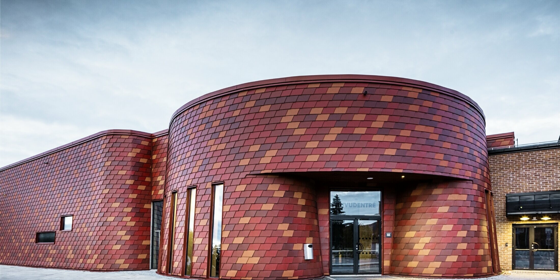 Patinoire à toit plat et façade incurvée ; la façade est revêtue de bardeaux de façade en aluminium de PREFA dans différentes nuances de rouge - rouge oxyde, rouge tuile, brun rouge, rouge brun